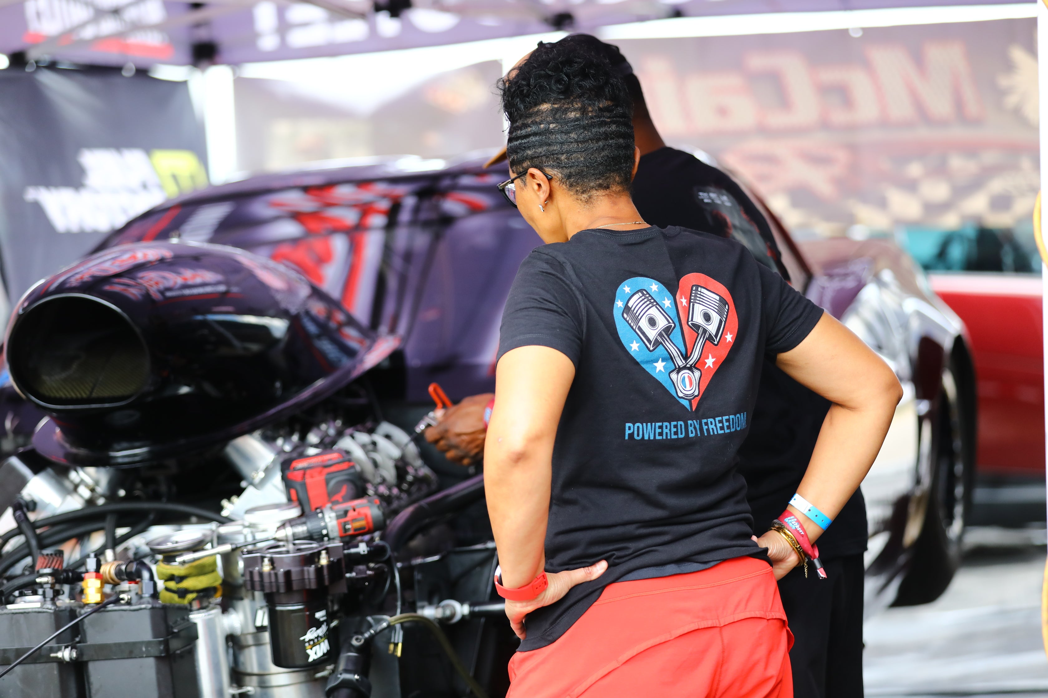 Person wearing a black t-shirt with a heart design and text, standing next to a car engine at an event.